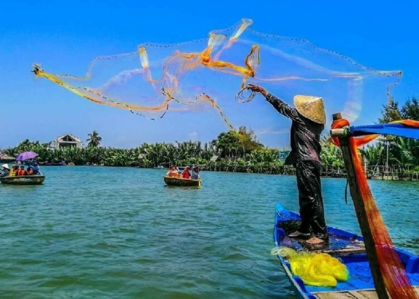 A fisherman in a conical hat stands on a wooden boat casting a large yellow throw net in Hoi An, Vietnam.